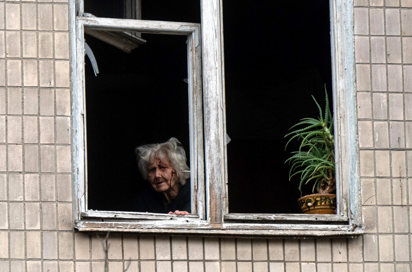Раненая пожилая женщина смотрит в разбитое окно пятиэтажного жилого дома, поврежденного российским беспилотником в Святошинском районе Киева